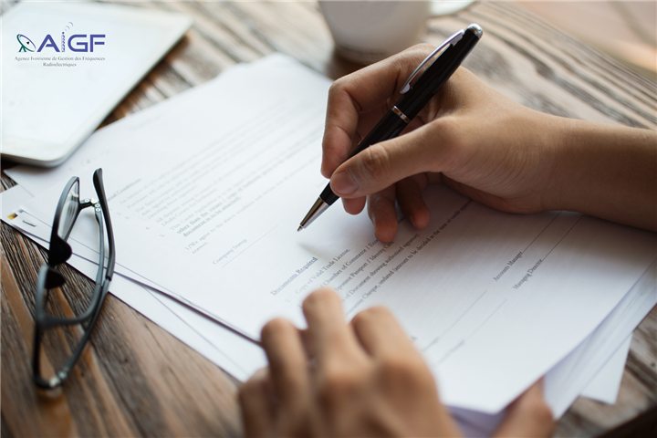 Deux mains signent un contrat posé sur un bureau en bois ; en haut à gauche, le logo de l'AIGF, Agence ivoirienne de gestion des fréquences radioélectriques.