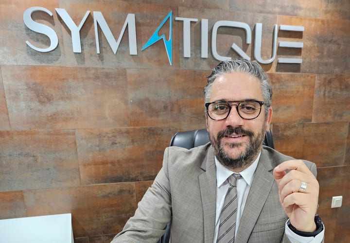 Photographie de l'interview : M. Amine Abdelkefi, en costume, assis à son bureau devant un ordinateur portable ; derrière lui, l'enseigne SYMATIQUE fixée au mur.