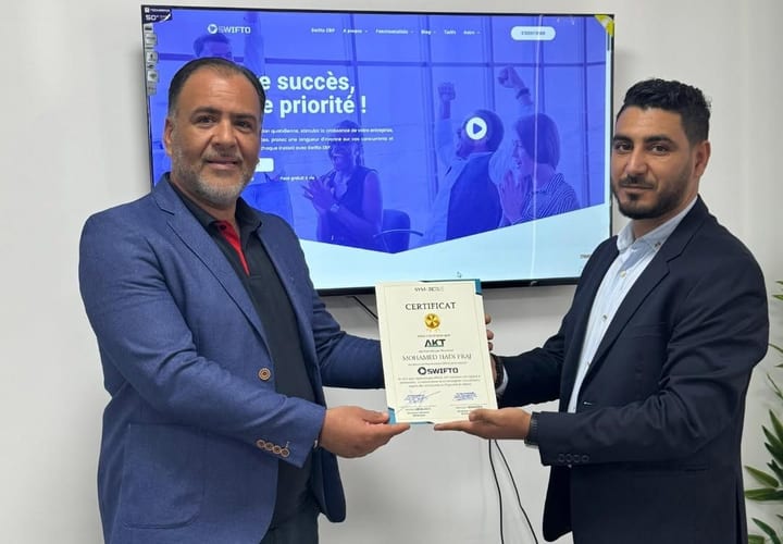 Photographie de l'annonce : deux hommes en costume tiennent ensemble un certificat au nom d'AKT Solutions, remis par SYMATIQUE ; derrière eux, un écran affiche le site de Swifto ERP.