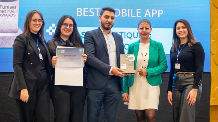 Photographie de la remise du prix : cinq personnes sur scène, l'une tenant un certificat de lauréat et une autre le trophée des Tunisia Digital Awards ; derrière elles, l'écran affiche « BEST MOBILE APP ».