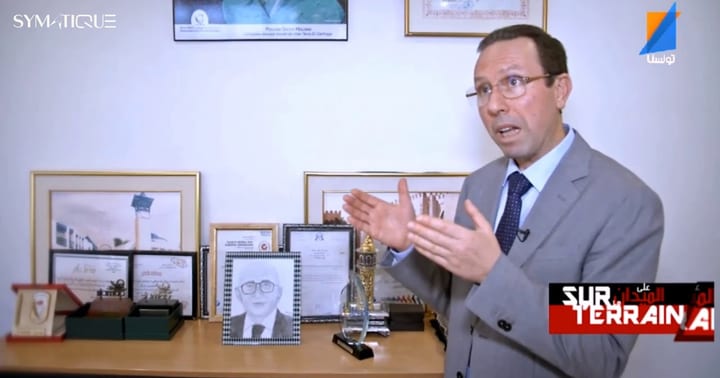 لقطة من الفيديو: السيد نوفل ساسي ببذلة رسمية يتحدّث في مكتبه أمام خزانة من الجوائز والشهادات المؤطَّرة؛ وفي أعلى اليسار شعار SYMATIQUE.