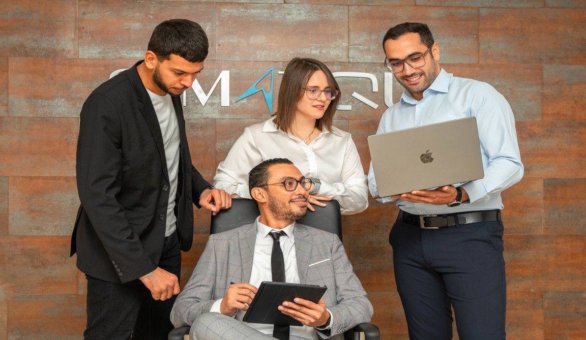 Cinq collaborateurs de SYMATIQUE côte à côte devant le mur de marque du siège, aux Berges du Lac à Tunis.