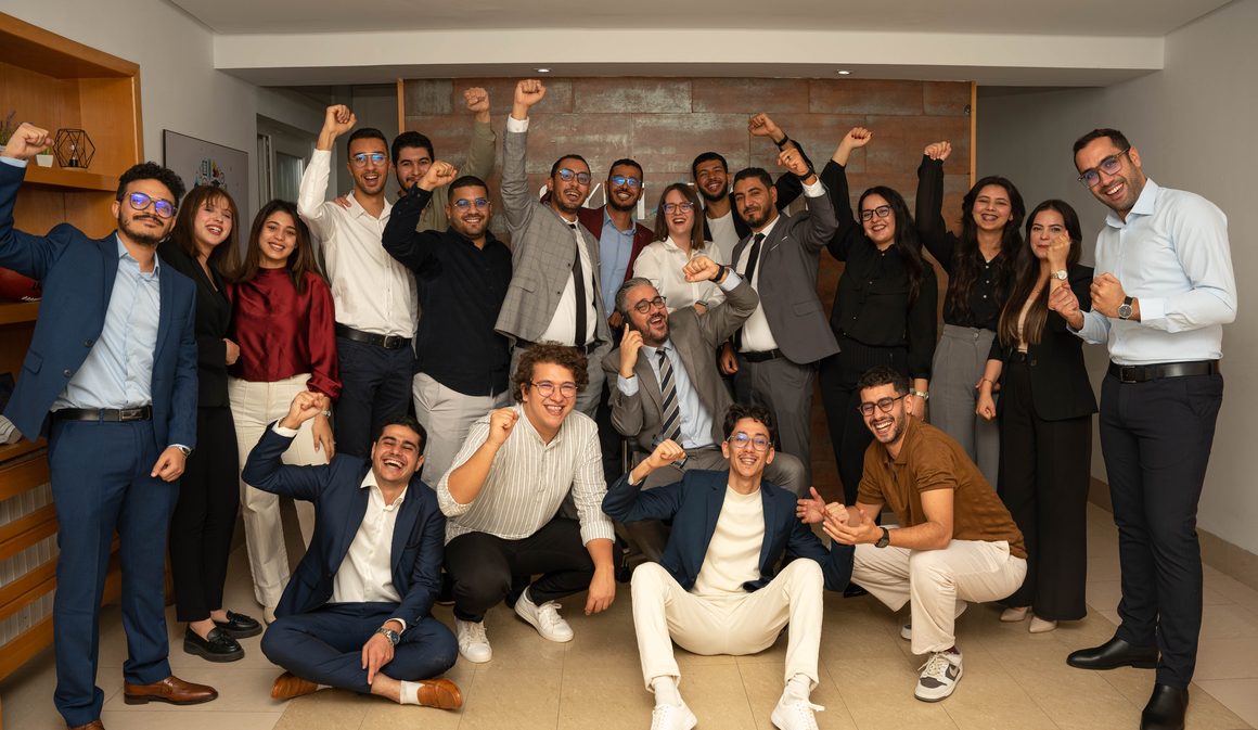 L'équipe de SYMATIQUE réunie dans les bureaux du siège devant le mur de marque, poings levés et sourires : certains debout, d'autres accroupis ou assis au sol.
